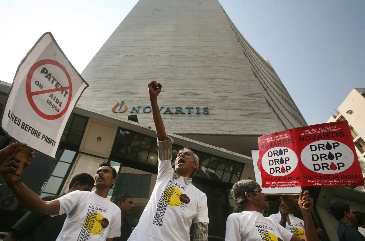 HIV-rights activists stand in front of a tall Novartis office building in Mumbai, holding signs that read “No patents on AIDS drugs” and “Drop the case,” during a street protest.